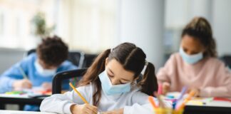 Girl In Mask Sitting At Desk Writing In Classroom