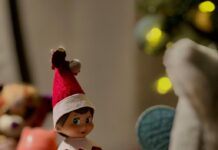 Stock photo of a small toy elf wearing a red shirt and red hat. He is sitting inside a Croc shoe with the paper note "All aboard the shoe train."