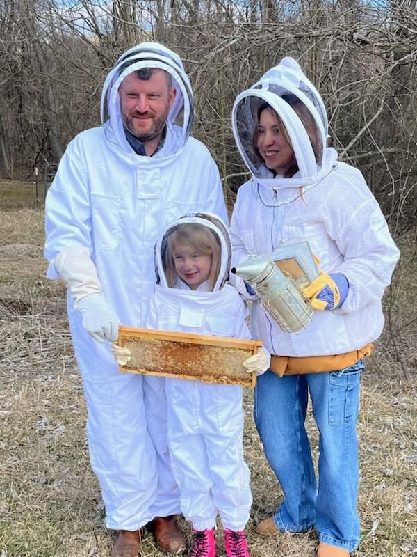 Two adults and a small child are dressed in white beekeeping uniforms and the child is holding a part of the hive framework