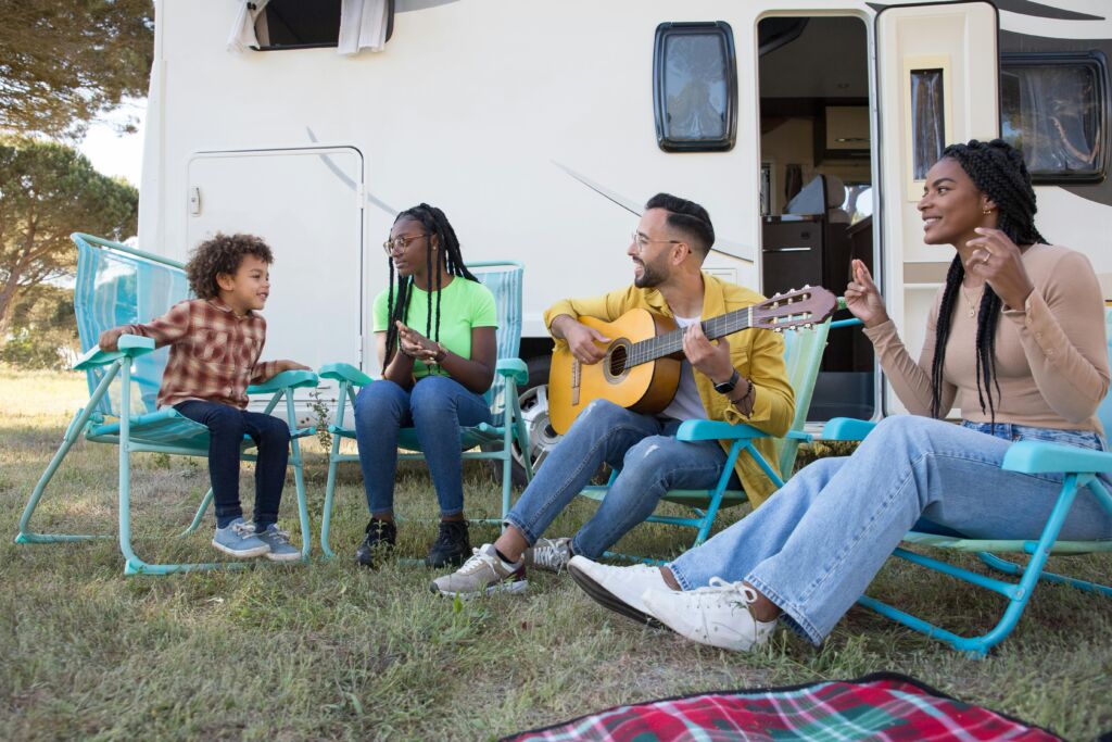 Three adults and a child sit in chairs outside their RV, with one man strumming a guitar
