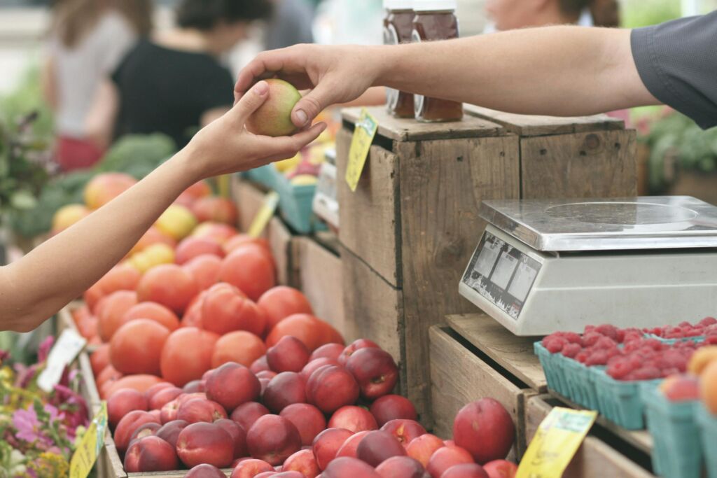 A vendor hands a peach or other piece of fruit to someone at a farmers market. The camera shows only their arms reaching out to make the exchange.