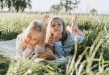 Two school-age girls are reading books out in a grassy field in the summertime.
