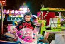 A boy sitting in a pink carnival ride car enjoys a night ride with lights in the background.