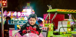 A boy sitting in a pink carnival ride car enjoys a night ride with lights in the background.