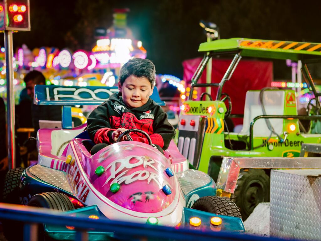 A boy sitting in a pink carnival ride car enjoys a night ride with lights in the background.